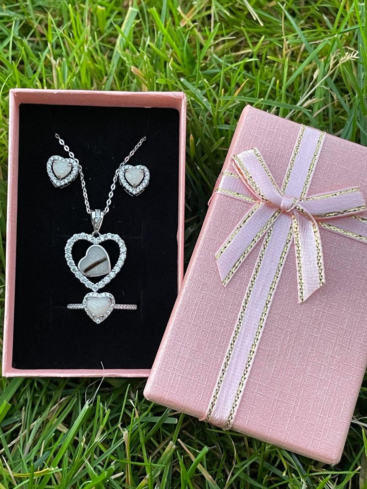 Silver heart-shaped breastmilk jewelry set with matching necklace, ring, and earrings displayed in a pink gift box on green grass.