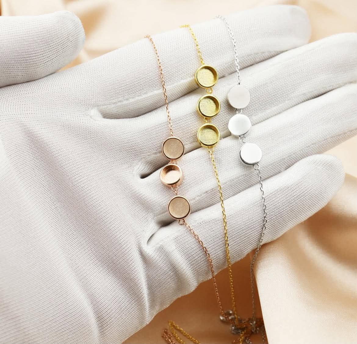 Three delicate disc-chain necklaces in gold, rose gold, and silver tones are displayed on a white gloved hand against a soft beige satin background.