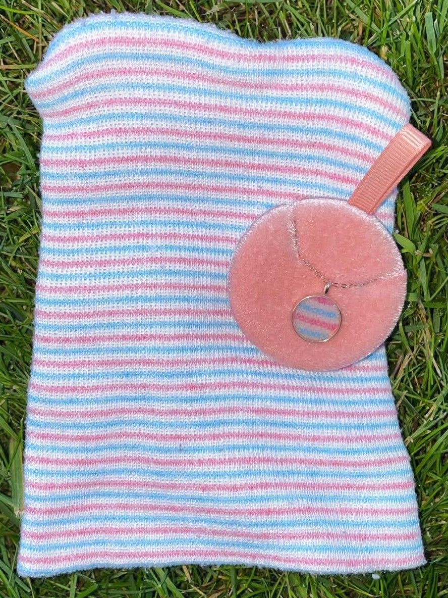 Round rose gold keepsake necklace containing a piece of a pink, blue, and white striped baby hat, displayed beside the original newborn hospitsl hat on green grass.