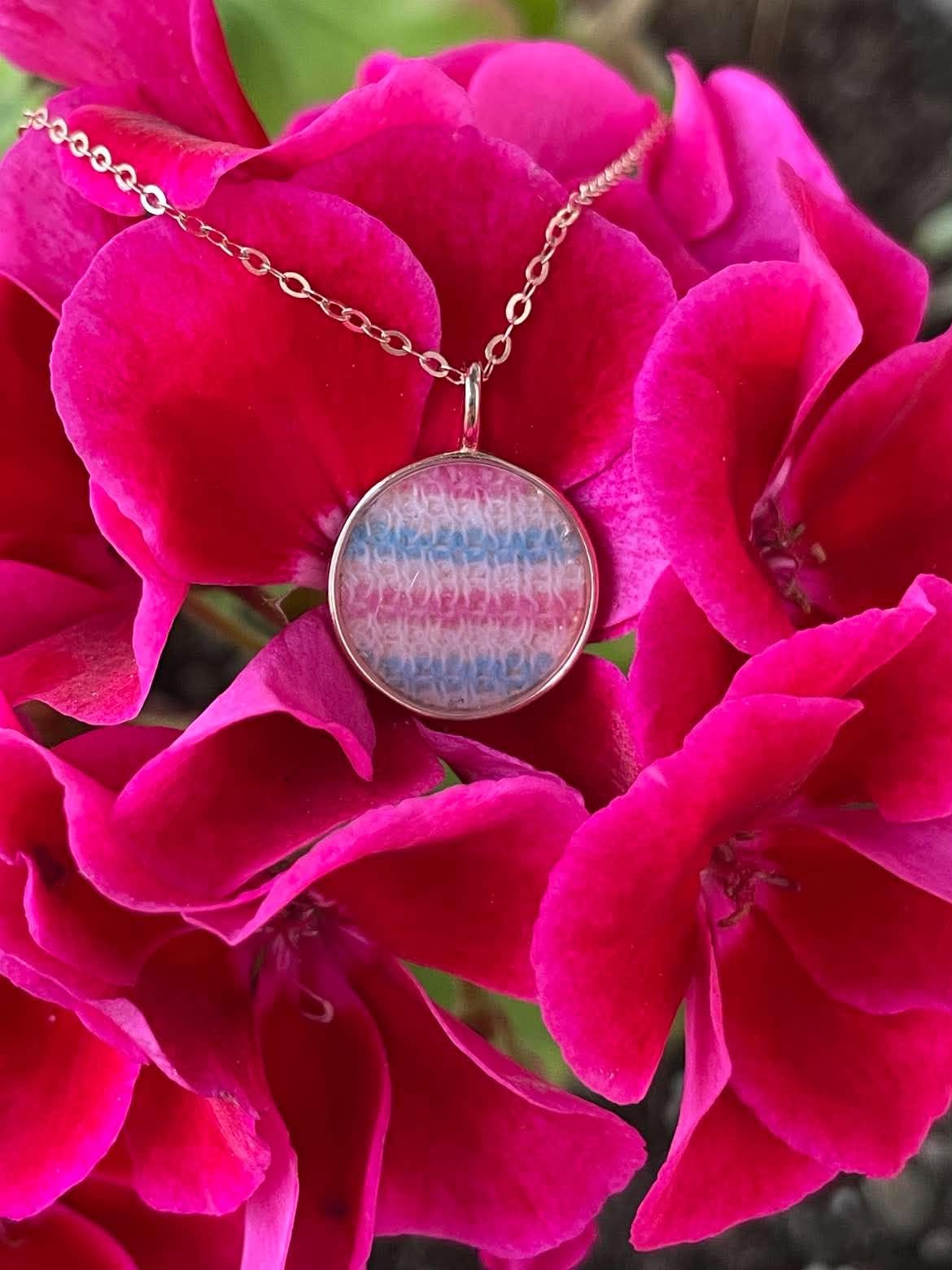 Round rose gold keepsake necklace containing a piece of a baby hat with pink, blue, and white knit fabric, displayed against bright pink flowers.