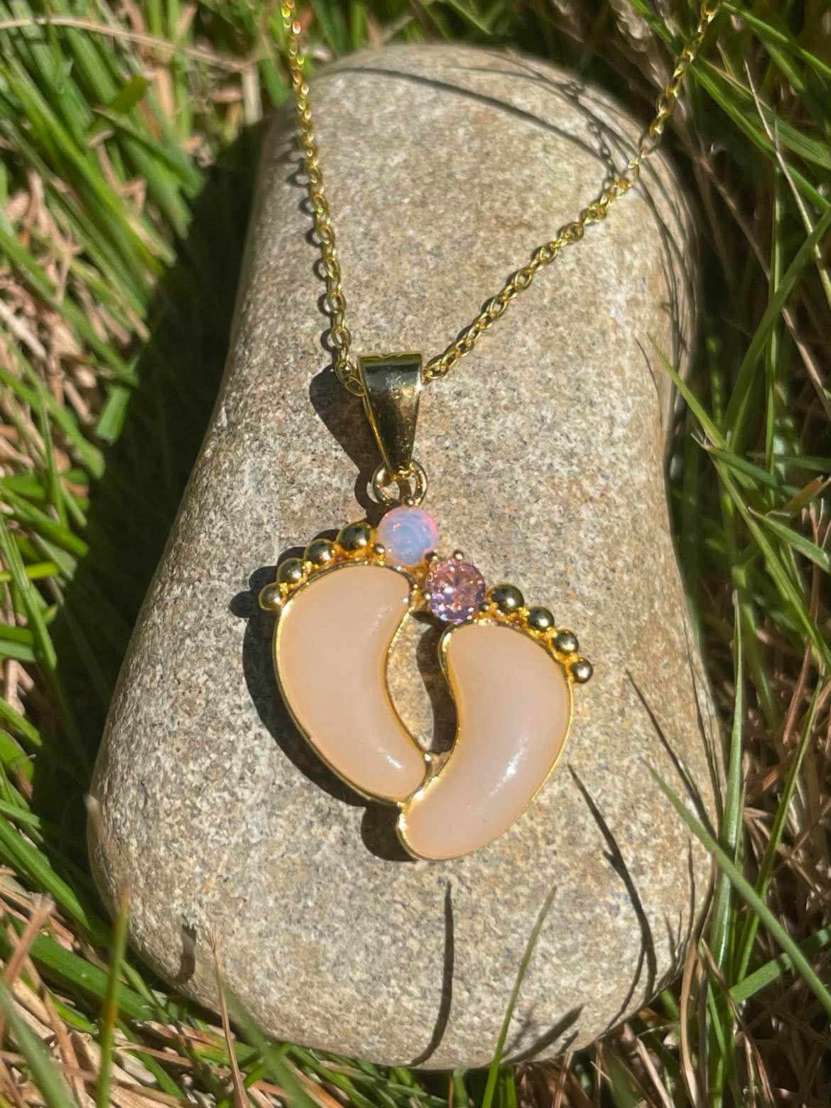 Close up of gold baby steps necklace with a feet design filled with breastmilk and has 2 birthstones attatched to the toes, moonstone and pink displayed on a rock in the sunlight.
