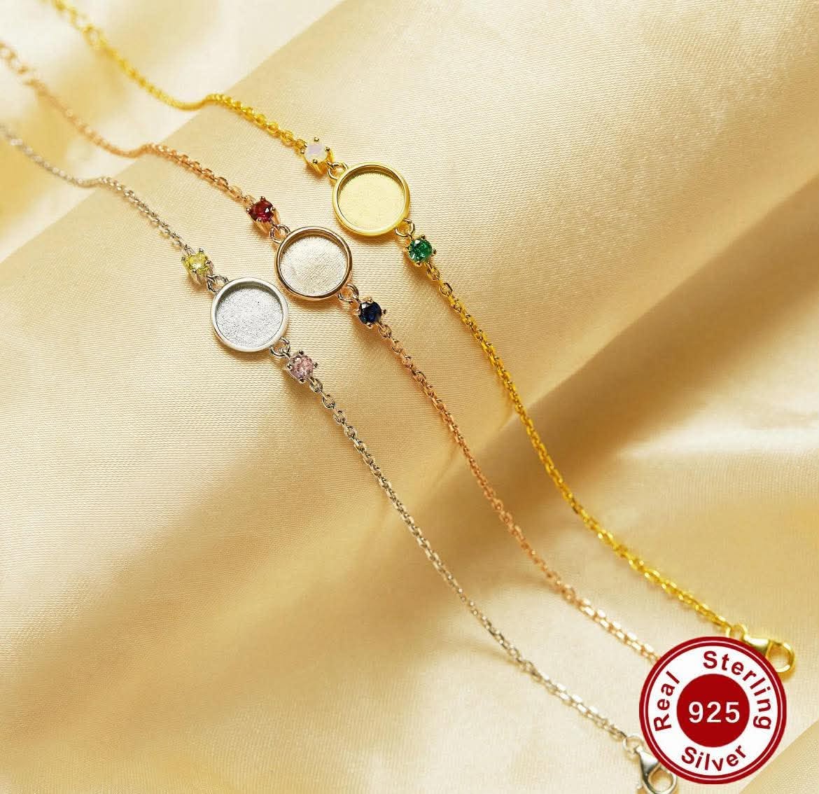 Three delicate chain bracelets in yellow gold, rose gold, and silver, each featuring a circular bezel setting with small colorful birthstones, displayed on a draped champagne satin fabric. A 'Real Sterling 925 Silver' badge is visible in the bottom right corner.