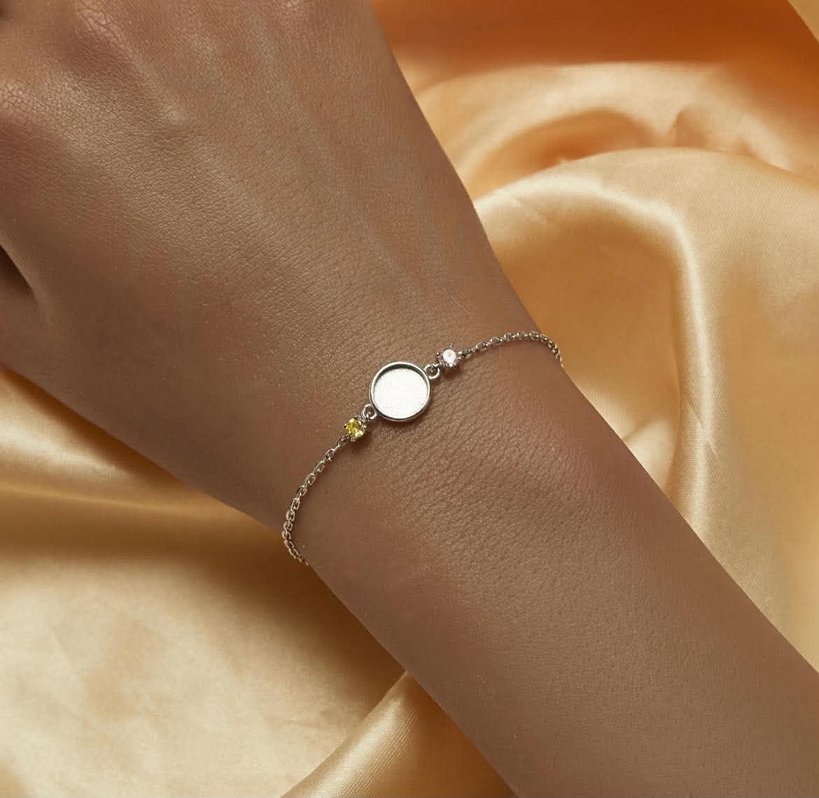 A delicate silver chain bracelet with a round bezel-set white center, a small pink birthstone, and a tiny yellow gemstone, shown worn on a wrist against a draped champagne silk background.