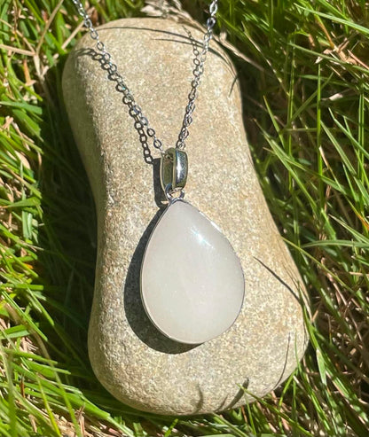 Silver teardrop-shaped breastmilk keepsake necklace with a soft white stone, displayed on a smooth rock surrounded by green grass in bright natural sunlight.