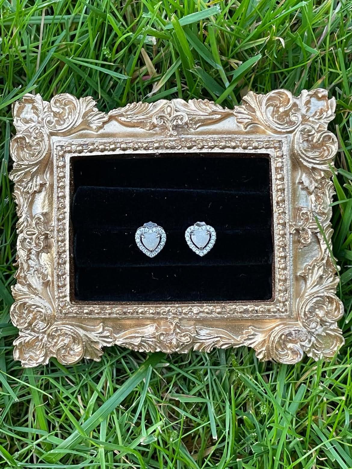 Silver heart-shaped breastmilk stud earrings with sparkling crystal halos displayed on black velvet inside an ornate gold frame on green grass.