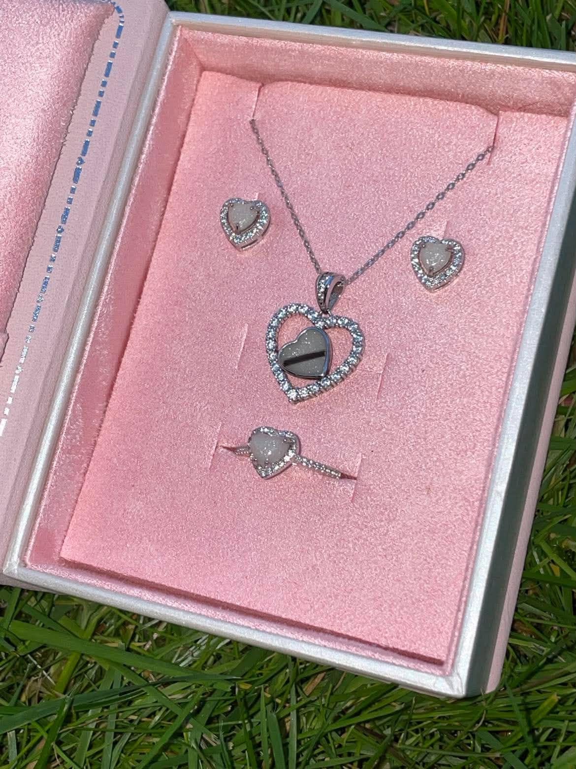 Silver heart-shaped breastmilk keepsake jewelry set with matching necklace, ring, and earrings displayed in a pink jewelry box on green grass.