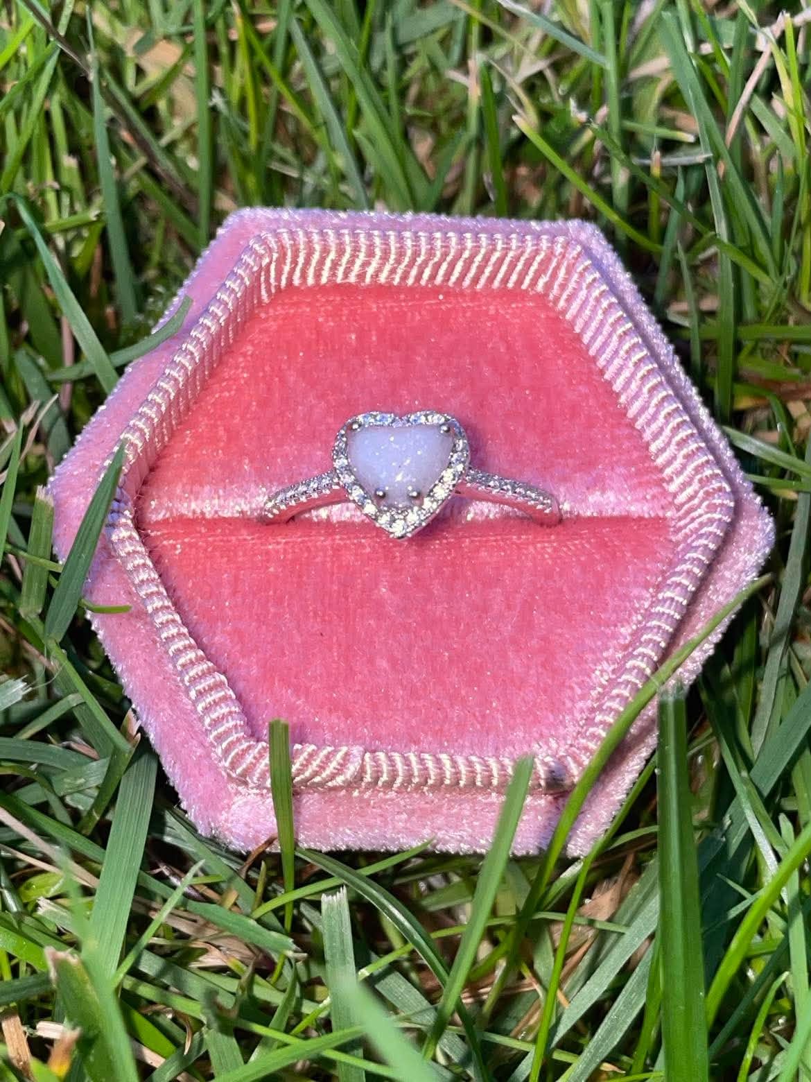 Silver heart-shaped breastmilk keepsake ring with sparkling crystal accents, displayed in a pink velvet ring box on green grass.