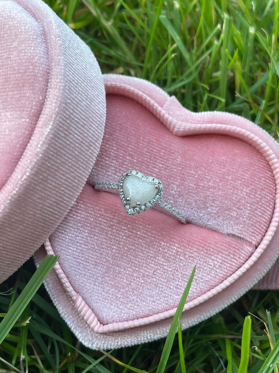 Silver heart-shaped breastmilk keepsake ring with sparkling crystal accents, displayed at an angle in an open pink velvet heart-shaped ring box on green grass.