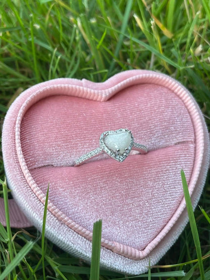 Silver heart-shaped breastmilk keepsake ring with sparkling crystal accents, displayed in a pink velvet heart-shaped ring box on green grass.