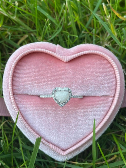 Silver heart-shaped breastmilk keepsake ring with sparkling crystal accents, displayed in a pink velvet heart-shaped ring box on green grass.