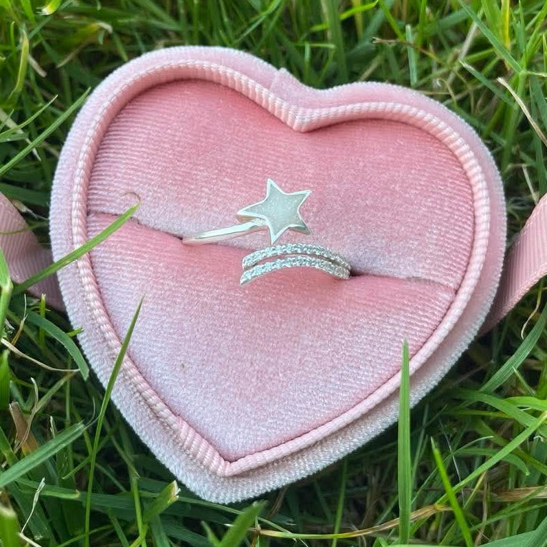 Silver star-shaped breastmilk ring with sparkling pavé bands displayed in a pink velvet heart-shaped ring box on green grass.