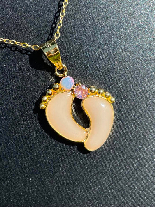 Close up of gold baby steps necklace with a feet design filled with breastmilk and has 2 birthstones attatched to the toes, moonstone and pink.