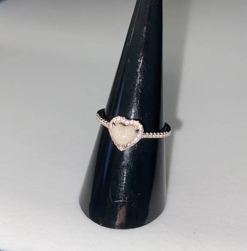 Silver heart-shaped breastmilk keepsake ring with a soft white center surrounded by sparkling stones, displayed on black cone.