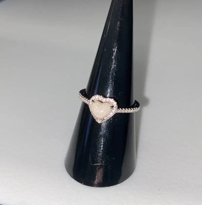 Silver heart-shaped breastmilk keepsake ring with a soft white center surrounded by sparkling stones, displayed on black cone.