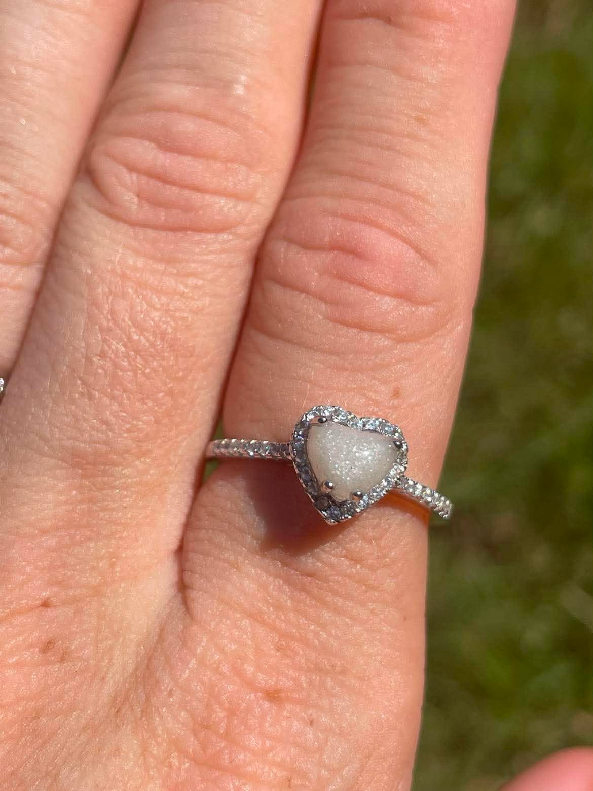Silver heart-shaped breastmilk keepsake ring with a soft white center surrounded by sparkling stones, displayed on finger.