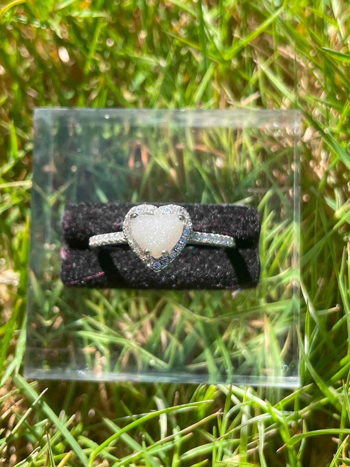 Silver heart-shaped breastmilk keepsake ring with a soft white center surrounded by sparkling stones, displayed in bright natural sunlight against green grass.