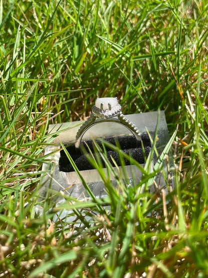 Silver heart-shaped breastmilk keepsake ring with a soft white center surrounded by sparkling stones, displayed in bright natural sunlight against green grass completly side view.