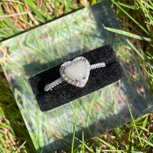 Silver heart-shaped breastmilk keepsake ring with a soft white center surrounded by sparkling stones, displayed in bright natural sunlight against green grass.