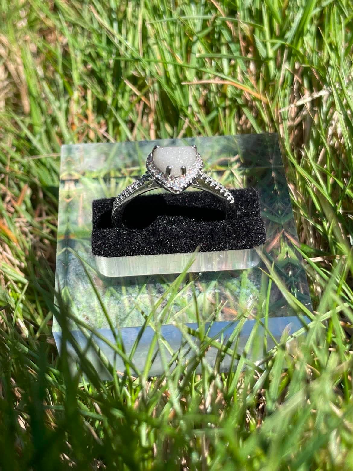Silver heart-shaped breastmilk keepsake ring with a soft white center surrounded by sparkling stones, displayed front side view in bright natural sunlight against green grass.