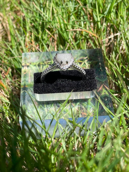 Silver heart-shaped breastmilk keepsake ring with a soft white center surrounded by sparkling stones, displayed front side view in bright natural sunlight against green grass.