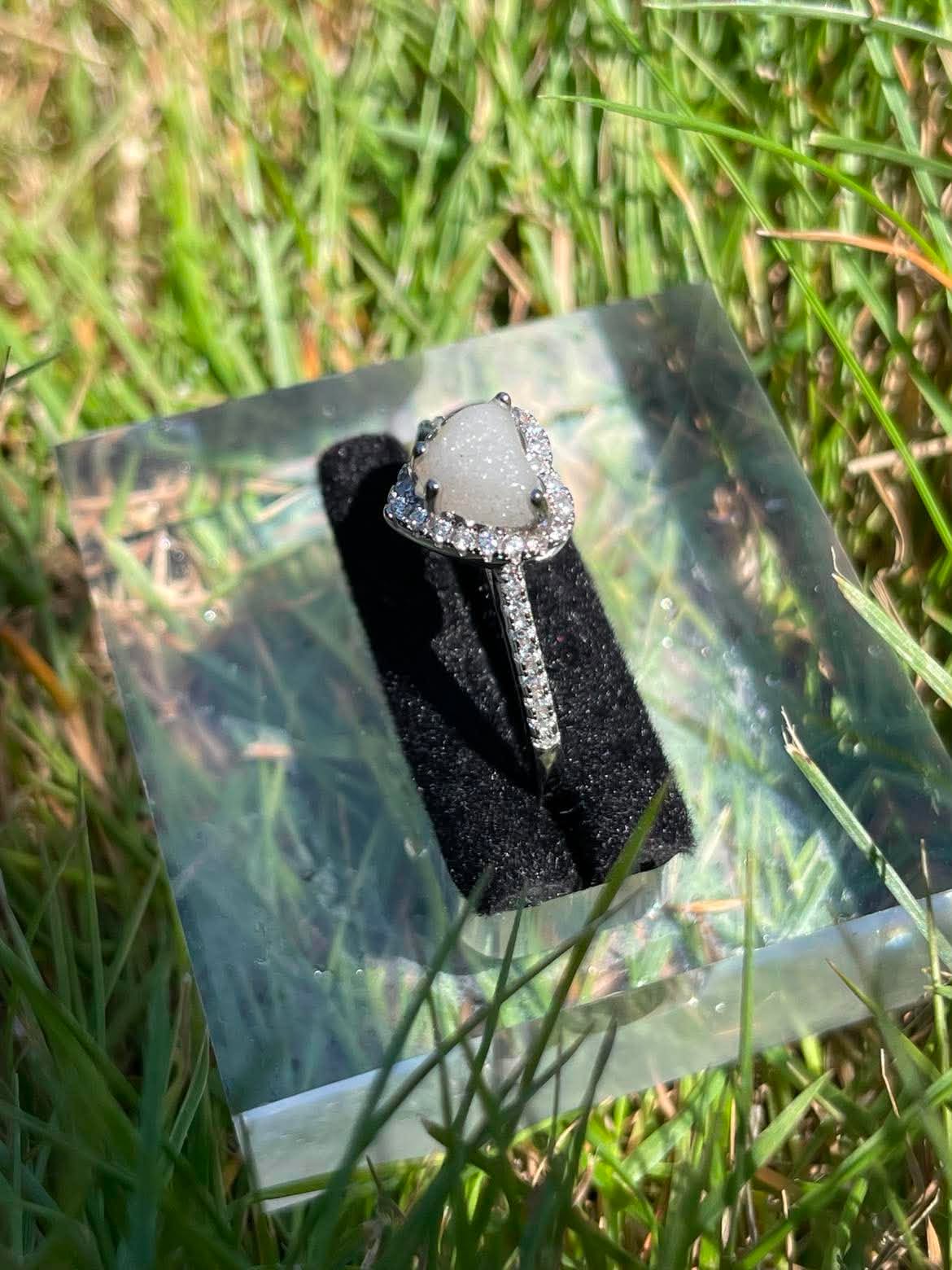 Silver heart-shaped breastmilk keepsake ring with a soft white center surrounded by sparkling stones, displayed in a side view in bright natural sunlight against green grass.