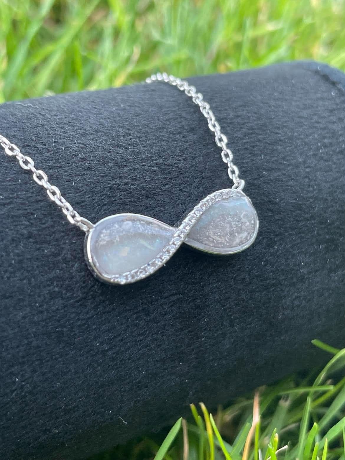 Silver infinity necklace featuring breastmilk and ashes keepsake stones with sparkling crystal accents, displayed on black velvet outdoors.