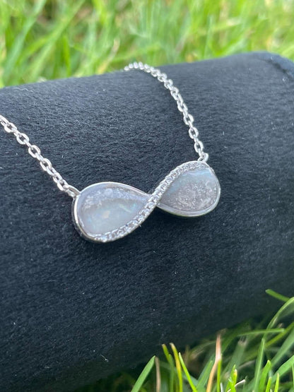 Silver infinity necklace featuring breastmilk and ashes keepsake stones with sparkling crystal accents, displayed on black velvet outdoors.