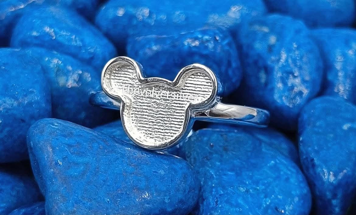 A silver Mickey Mouse-shaped ring with a hollow face and a polished band, resting on a bed of smooth, glossy blue stones.