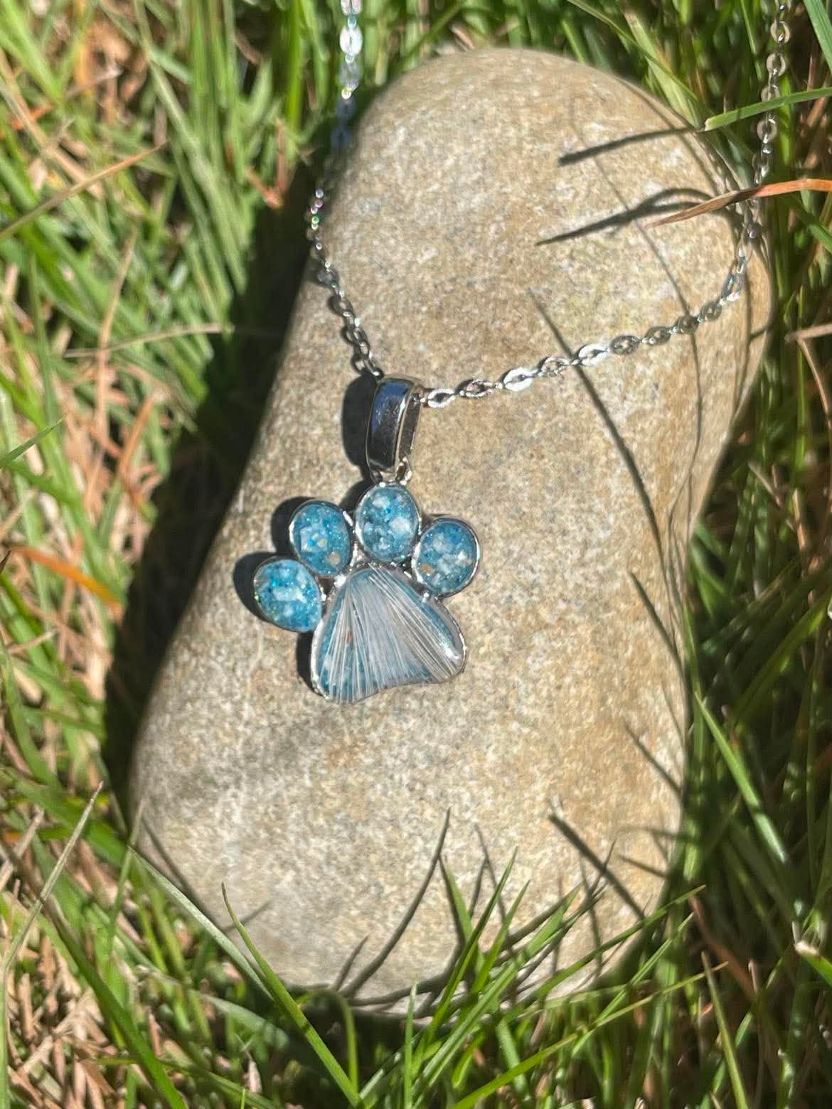 Silver pawprint necklace filled with blue color and ashes and fur from dog, displayed on rock in sunlight.