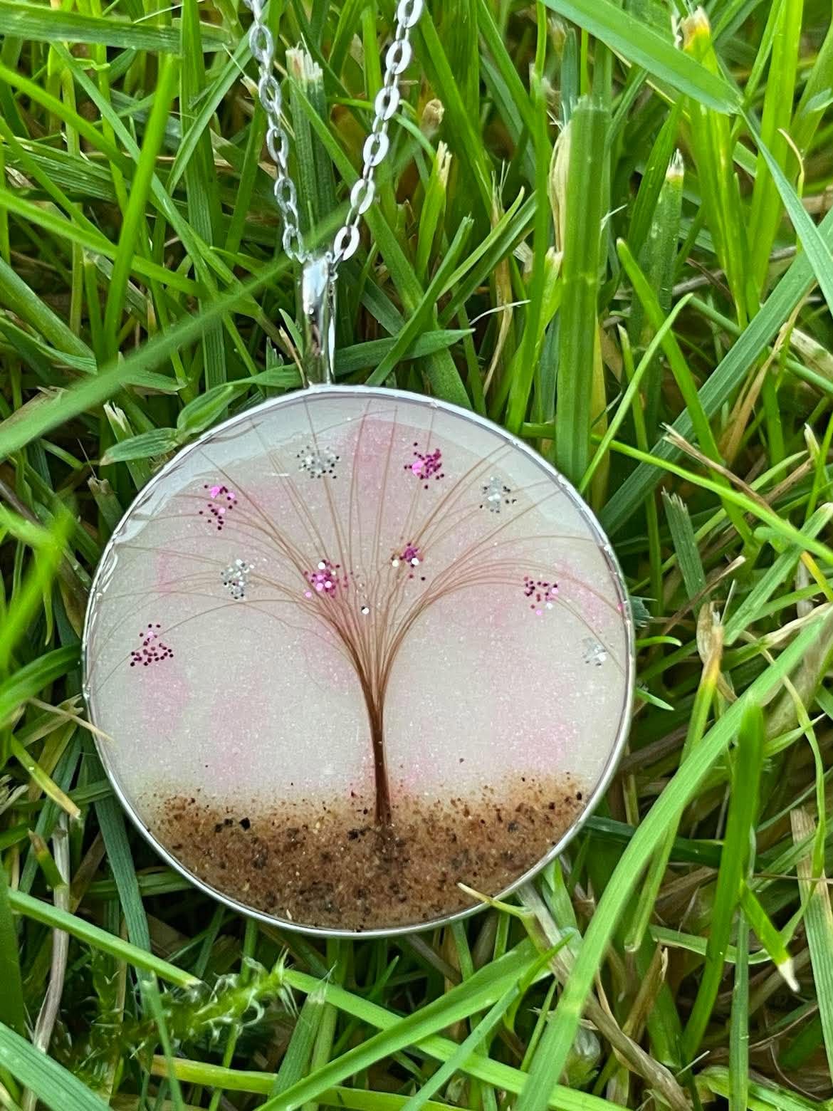 Round silver keepsake necklace featuring a tree design made with hair, preserved placenta, and pink glitter accents, displayed on green grass.