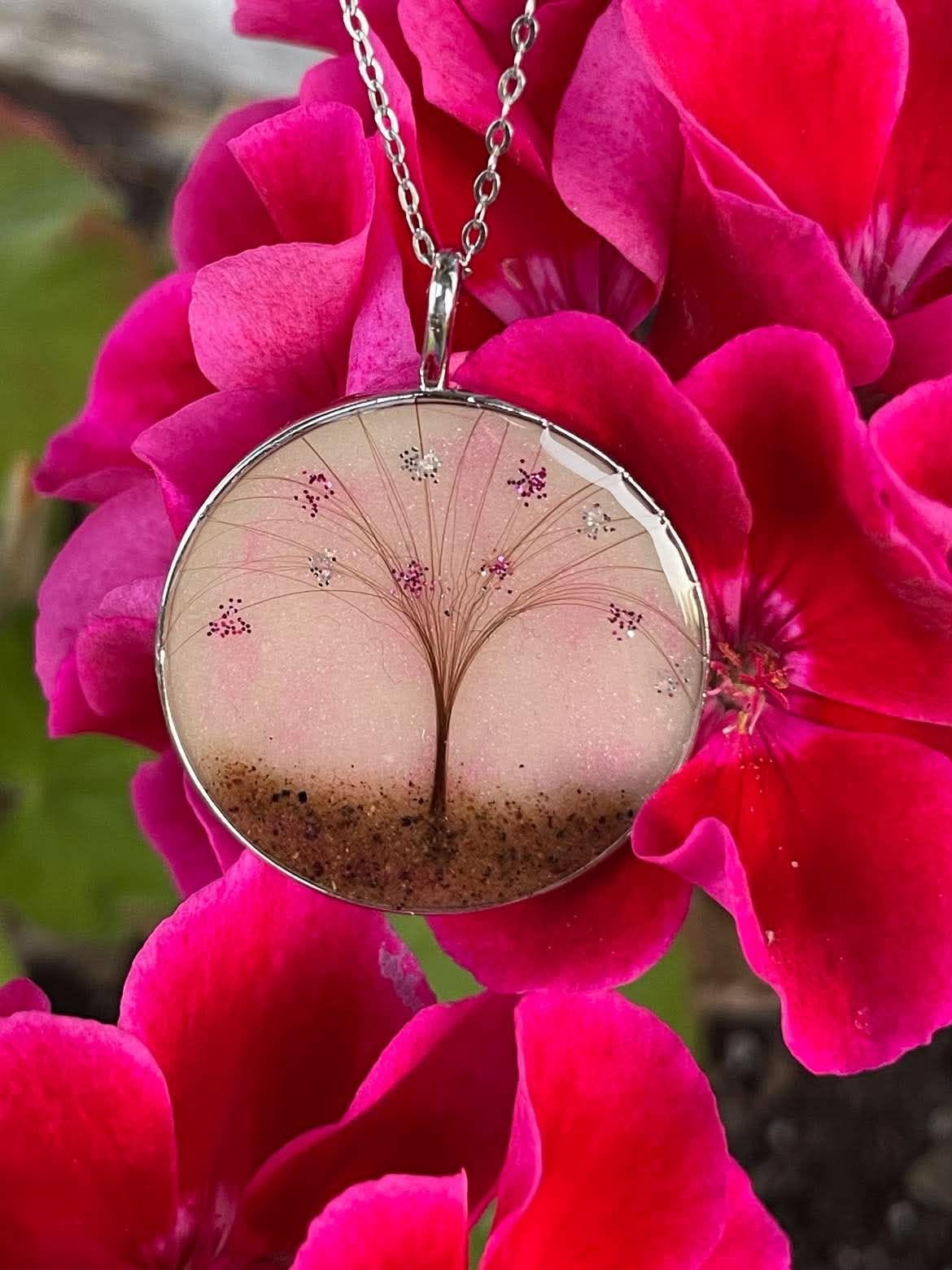 Round silver keepsake necklace featuring a tree design made with hair, preserved placenta, and pink glitter accents, displayed against bright pink flowers.