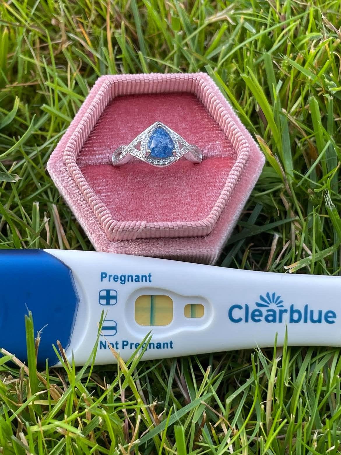 Silver teardrop-shaped pregnancy test keepsake ring with a blue center, displayed in a pink velvet ring box beside the original positive Clearblue pregnancy test on green grass.