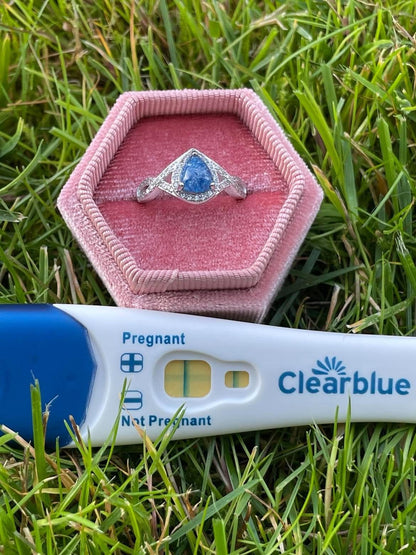 Silver teardrop-shaped pregnancy test keepsake ring with a blue center, displayed in a pink velvet ring box beside the original positive Clearblue pregnancy test on green grass.