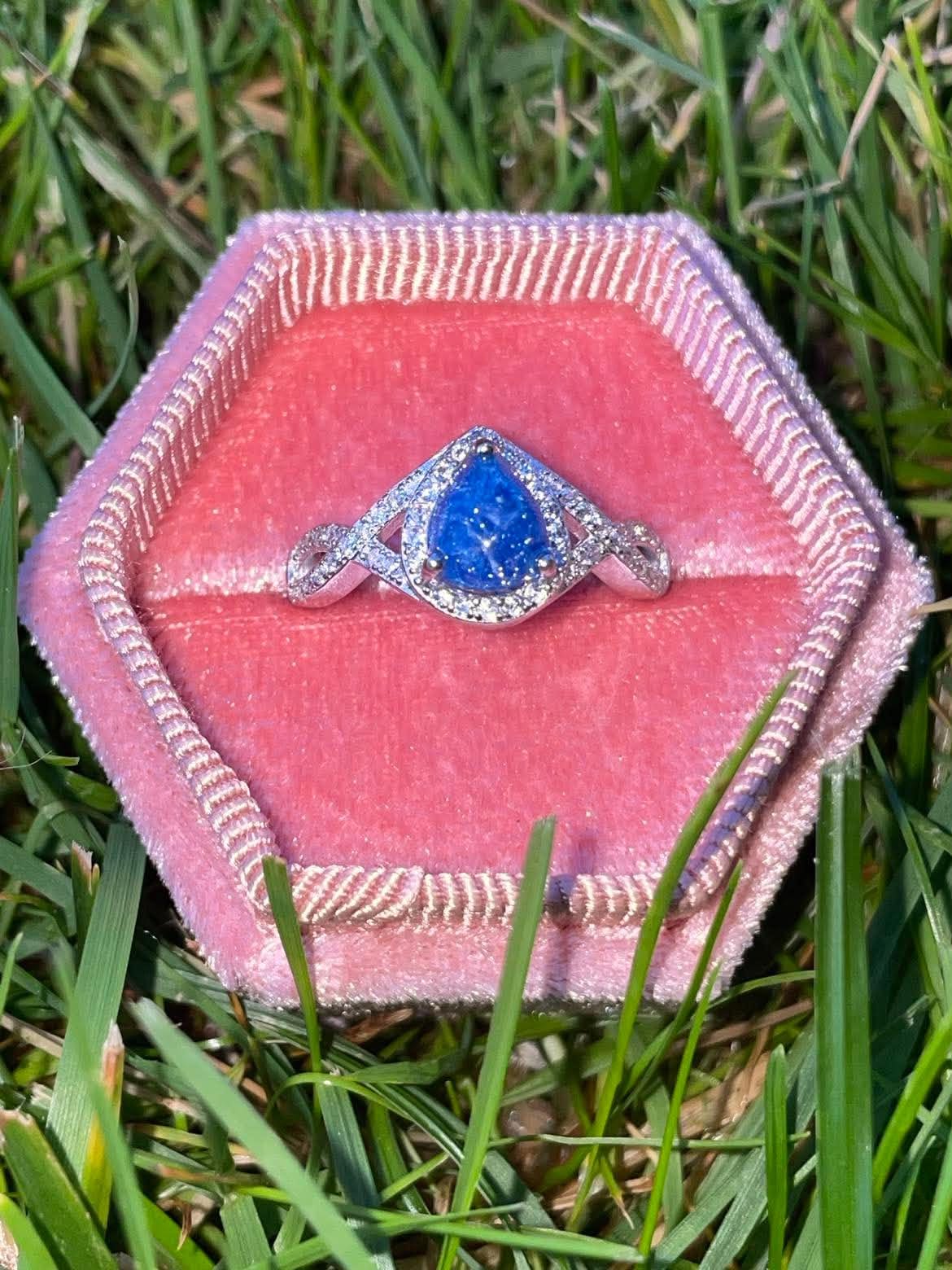 Silver teardrop-shaped pregnancy test keepsake ring with a vivid blue center and sparkling crystal accents, displayed in a pink velvet ring box on green grass.