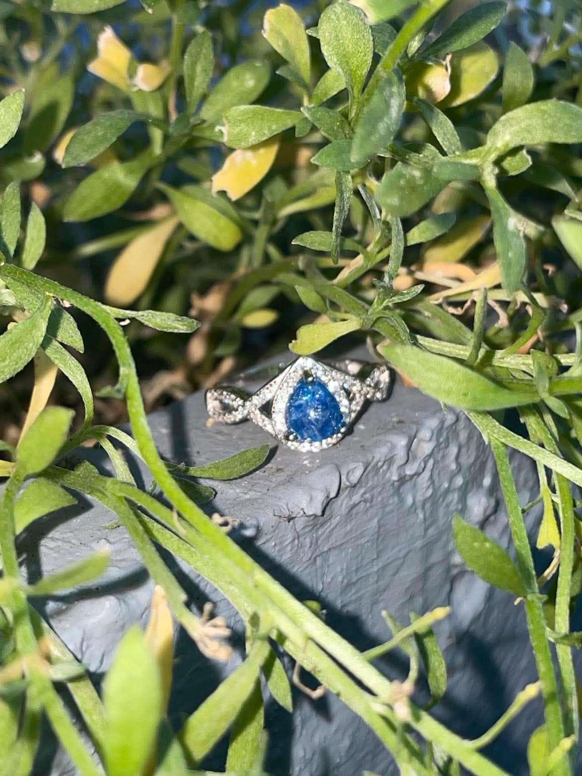 Silver teardrop-shaped pregnancy test keepsake ring with a vivid blue center and sparkling crystal accents, displayed outdoors among green leaves.