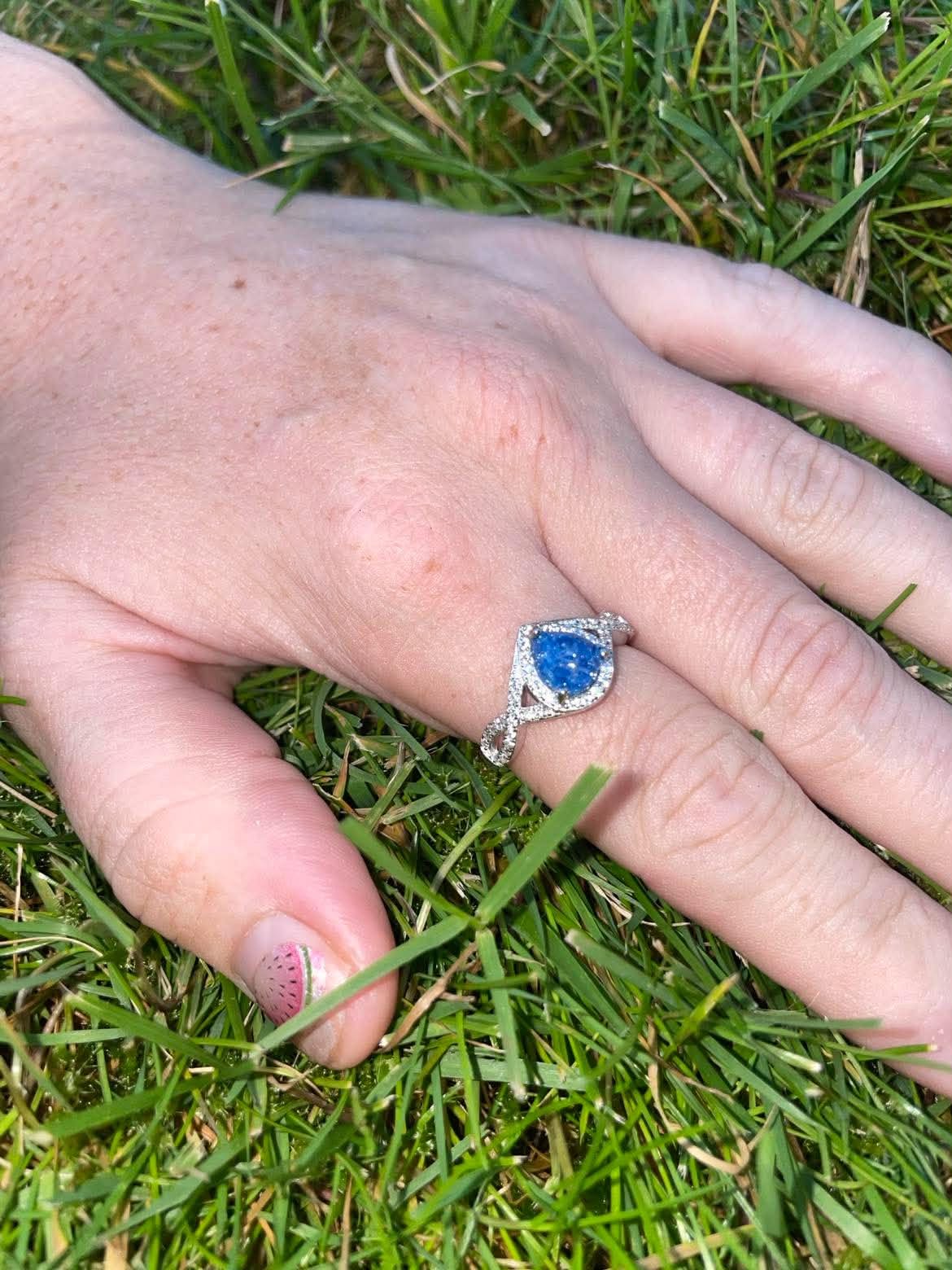 Silver teardrop-shaped pregnancy test keepsake ring with a bright blue center and sparkling crystal accents, worn on a hand resting in green grass.