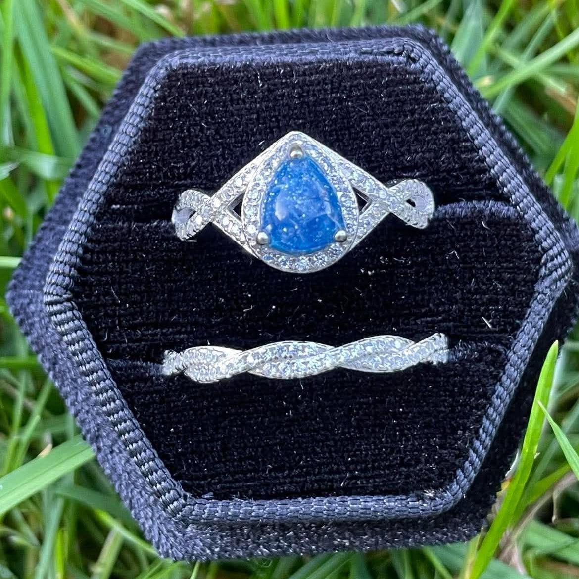 Silver teardrop-shaped pregnancy test keepsake ring with a bright blue center and sparkling crystal accents, paired with a matching twisted stacker ring in a black velvet ring box on green grass.
